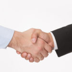 Close up of a handshake between a man in a black suit and a man in a blue button-up against a white background