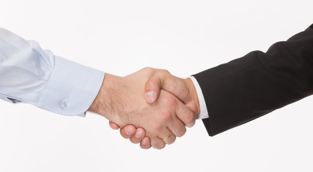 Close up of a handshake between a man in a black suit and a man in a blue button-up against a white background