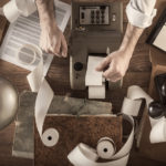 Man's forearms on a brown desk with a tax machine, antique phone, various papers, a lamp, a cigar, and a pen on it