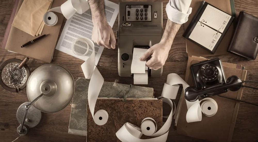 Man's forearms on a brown desk with a tax machine, antique phone, various papers, a lamp, a cigar, and a pen on it