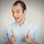 Man in a blue button-up shirt, smiling with his mouth open and making finger guns with his hands