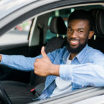 Man with a beard in a blue button up and white undershirt sitting in driver's seat of car giving thumbs up