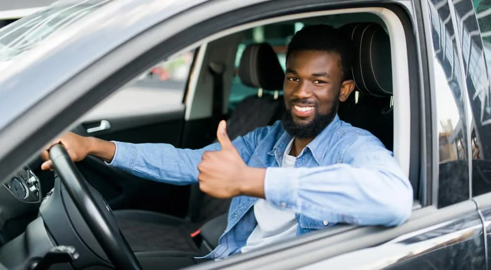 Man with a beard in a blue button up and white undershirt sitting in driver's seat of car giving thumbs up