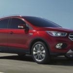 A red 2018 Ford Escape driving on a grassy road against a clear blue sky