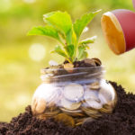 Watering can tilted toward a jar of change that is sticking out of the dark dirt ground with a green plant sticking out