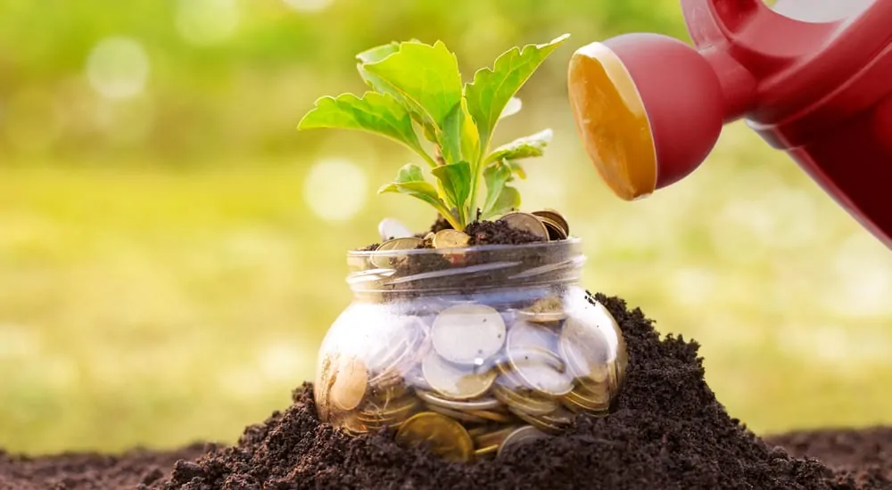 Watering can tilted toward a jar of change that is sticking out of the dark dirt ground with a green plant sticking out