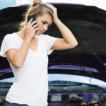Woman in a white t-shirt on the phone with hand on head in front of car with the hood open