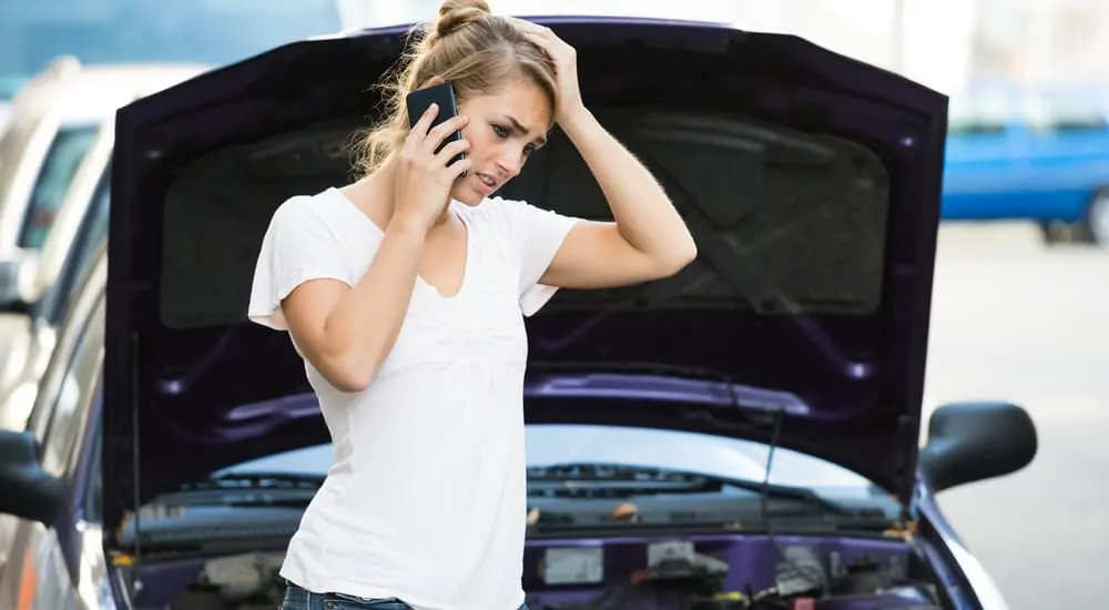 Woman in a white t-shirt on the phone with hand on head in front of car with the hood open