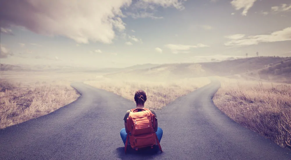 Woman in blue jeans and a t-shirt sitting toward a crossroads with an orange backpack