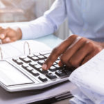 Man in a blue shirt at a desk using a calculator with a stack of receipts