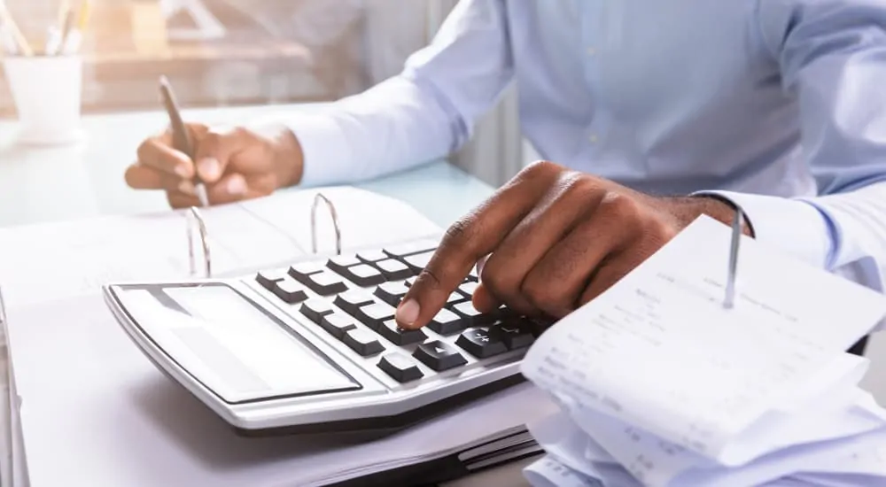 Man in a blue shirt at a desk using a calculator with a stack of receipts