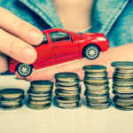 Woman in a blue shirt holding a red toy car above six increasingly taller stacks of coins