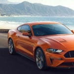 An orange Ford Mustang drives down a costal highway