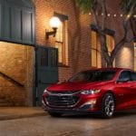 Red 2019 Chevrolet Malibu in front of brick building on tree lined street with lights across road