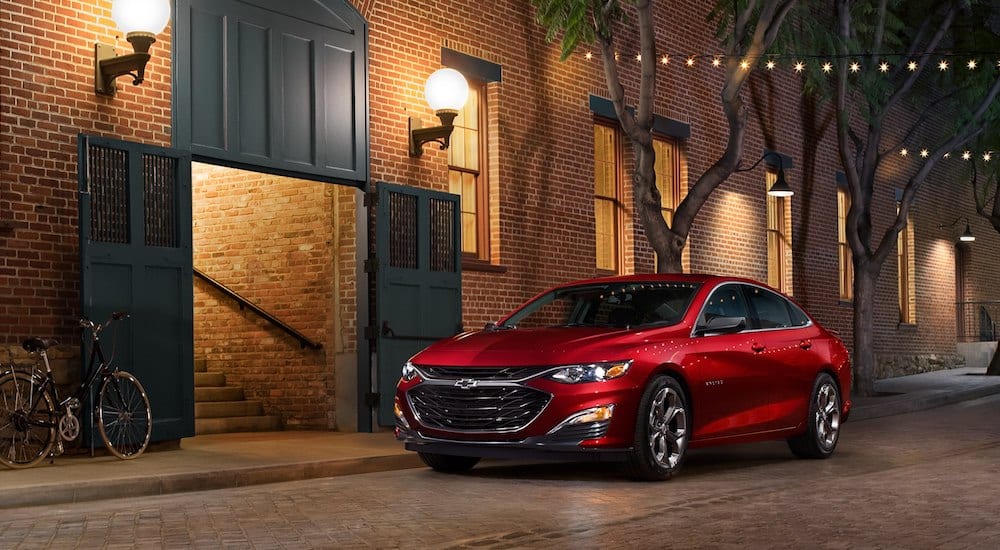 Red 2019 Chevrolet Malibu in front of brick building on tree lined street with lights across road