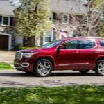 A red 2019 GMC Acadia driving in front of a stone house with trees flowering in back