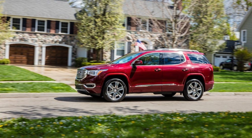 A red 2019 GMC Acadia driving in front of a stone house with trees flowering in back