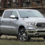 A silver 2020 Ram 1500 is parked in a grassy field with a rusty bridge in the background.