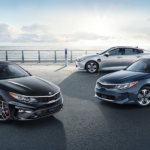 A black and a grey 2019 KIA Optima are parked in front of a silver plug in Optima with the ocean behind them.