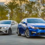A silver and a blue 2019 Kia Optima are parked in front of palm trees.