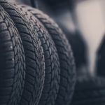 A closeup of tires in a tire shop is shown with an out of focus mechanic in the background.