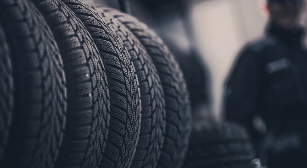 A closeup of tires in a tire shop is shown with an out of focus mechanic in the background.