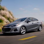 A silver 2019 Chevy Cruze is driving around a corner with a city view behind it.