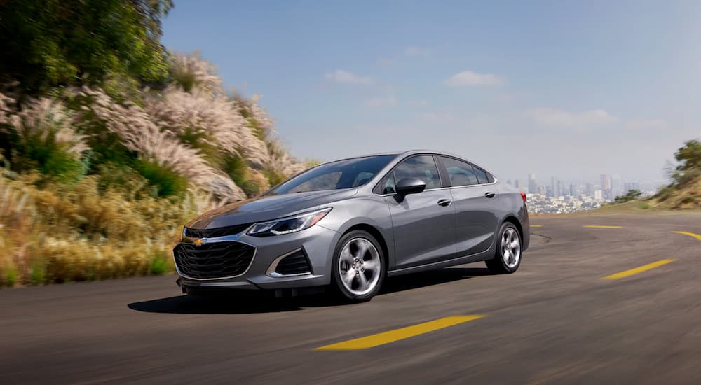 A silver 2019 Chevy Cruze is driving around a corner with a city view behind it.