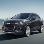 A grey 2019 Chevy Trax is driving with a city skyline and water behind it.