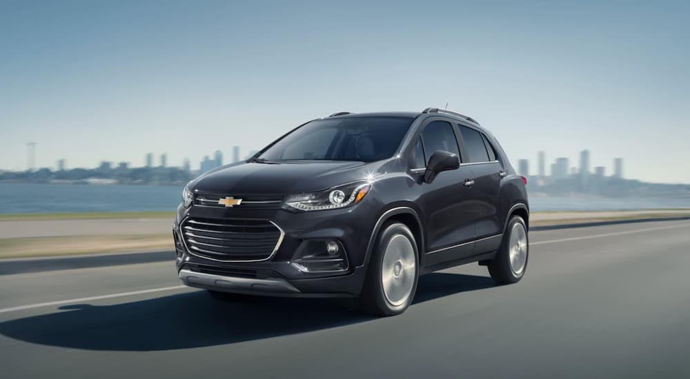 A grey 2019 Chevy Trax is driving with a city skyline and water behind it.