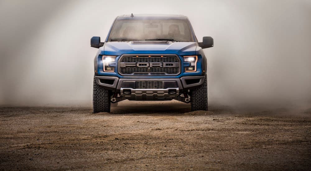 A blue 2019 Ford Raptor is driving on dirt with a dust cloud behind it.