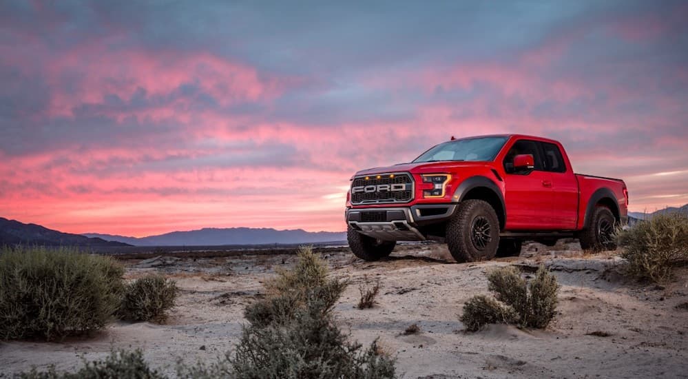 A red 2019 Ford Raptor, which wins when comparing the 2019 Ford Raptor vs 2019 Chevy Silverado Trail Boss, is parked on dirt with a sunset behind it. 