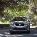 A silver 2020 Buick Envision is driving on a tree lined neighborhood road.