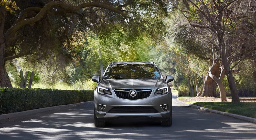 A silver 2020 Buick Envision is driving on a tree lined neighborhood road.