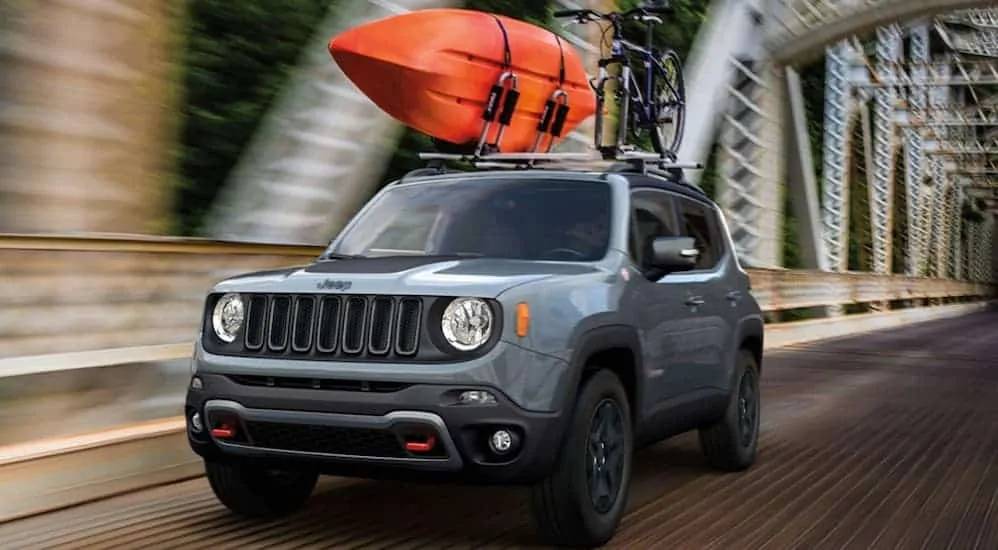 A blue 2020 Jeep Renegade is driving over a bridge with kayaks loaded on the roof racks.