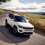 A white 2019 Jeep Compass is driving on a narrow, curvy road after leaving a Jeep dealership.