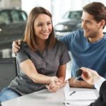 A car salesman is handing a couple the keys to the used car they purchased.