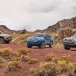 Three 2017 Toyota Tacoma's, a blue, brown, and grey colored, are parked on a dirt trail in the desert.
