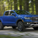 A blue Ford Ranger FX2 driving down a dirt road in a shadowy forest.