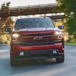 A red 2020 Chevy Silverado 1500 is driving on a tree lined road while facing forward.