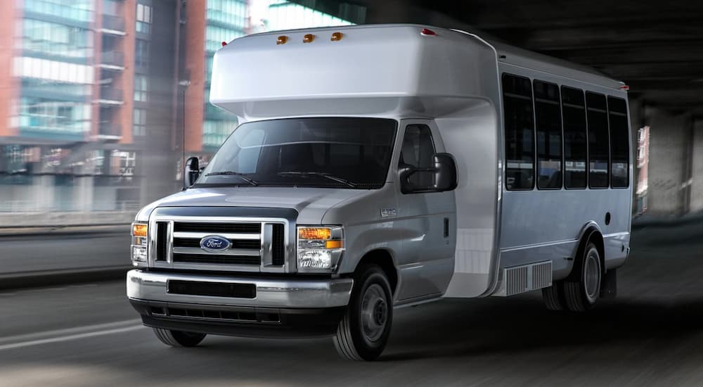 A white 2021 Ford E-Series with a bus upfit is driving on a city street at night.