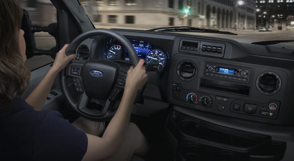 A driver is behind the wheel of one of the 2021 Ford commercial vehicles, an E-Series, on a road at night.