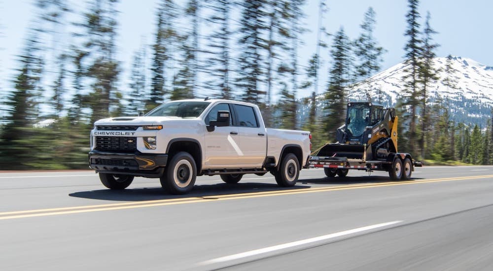 A white 2020 Chevy Silverado 2500HD diesel is towing construction equipment in front of evergreen trees.