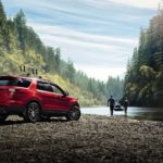 A red 2016 Ford Explorer is parked on a rocky shore while a couple brings a kayak to the water.