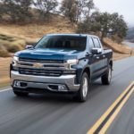 A blue 2020 Chevy Silverado 1500 is driving on a tree lined road.