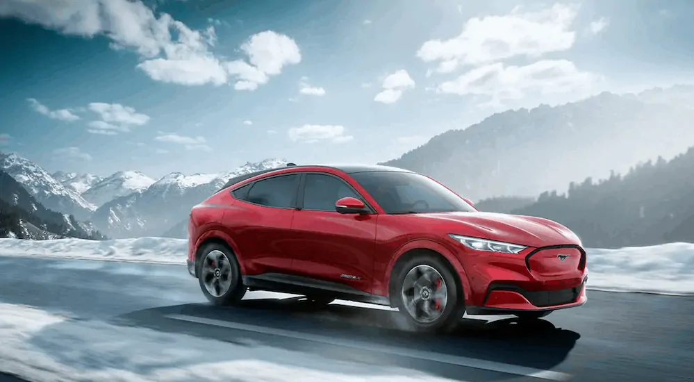 A red 2021 Ford Mustang Mach-E is driving on a road with snow covered mountains in the distance.