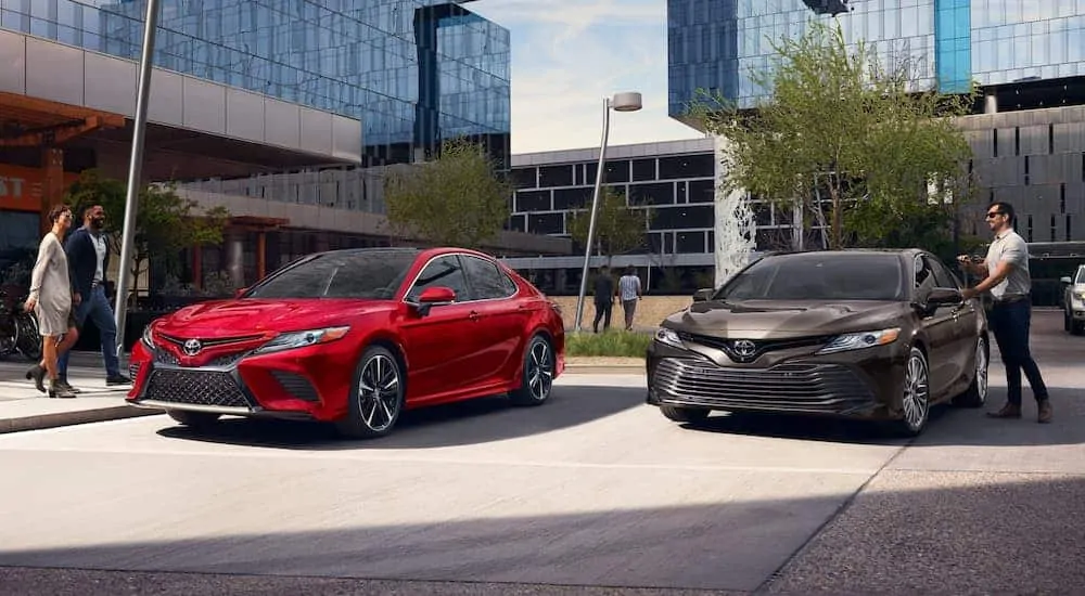 A red 2019 Toyota Camry is parked next to a dark grey one in front of buildings with lots of windows.