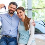 A husband has his arm around his wife and his new keys in his hand while they lean on a used car for sale.