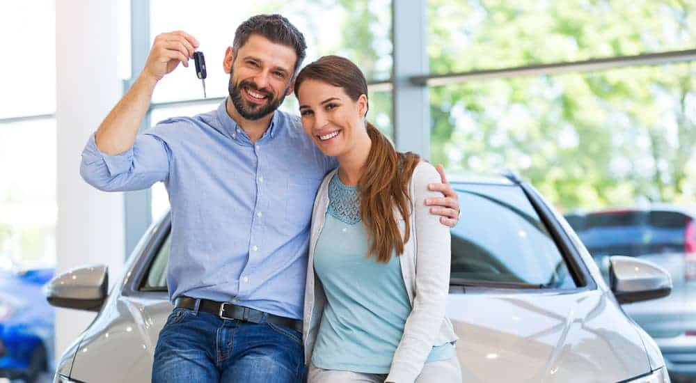 A husband has his arm around his wife and his new keys in his hand while they lean on a used car for sale.