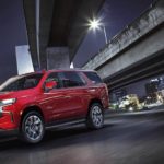 A red 2021 Chevy Tahoe is driving on a highway with a night time city skyline in the distance.